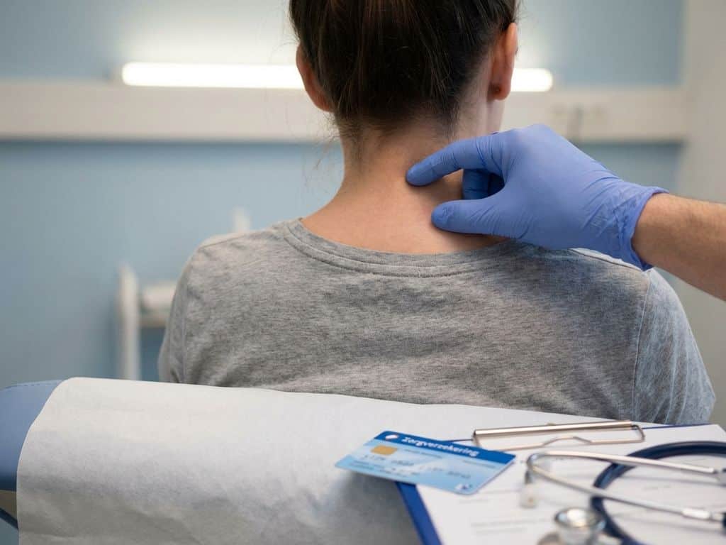 Zachte hand drukt op nekwervels tijdens fysiotherapie, zorgpas op onderzoekstafel in klinische omgeving